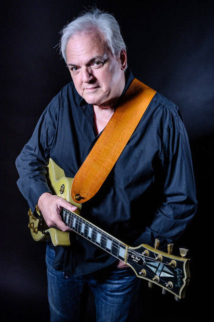 Mann mit grauen Haaren trägt schwarze Hemd und hält eine E-Gitarre mit braunem Gitarrengurt vor dunklem Hintergrund