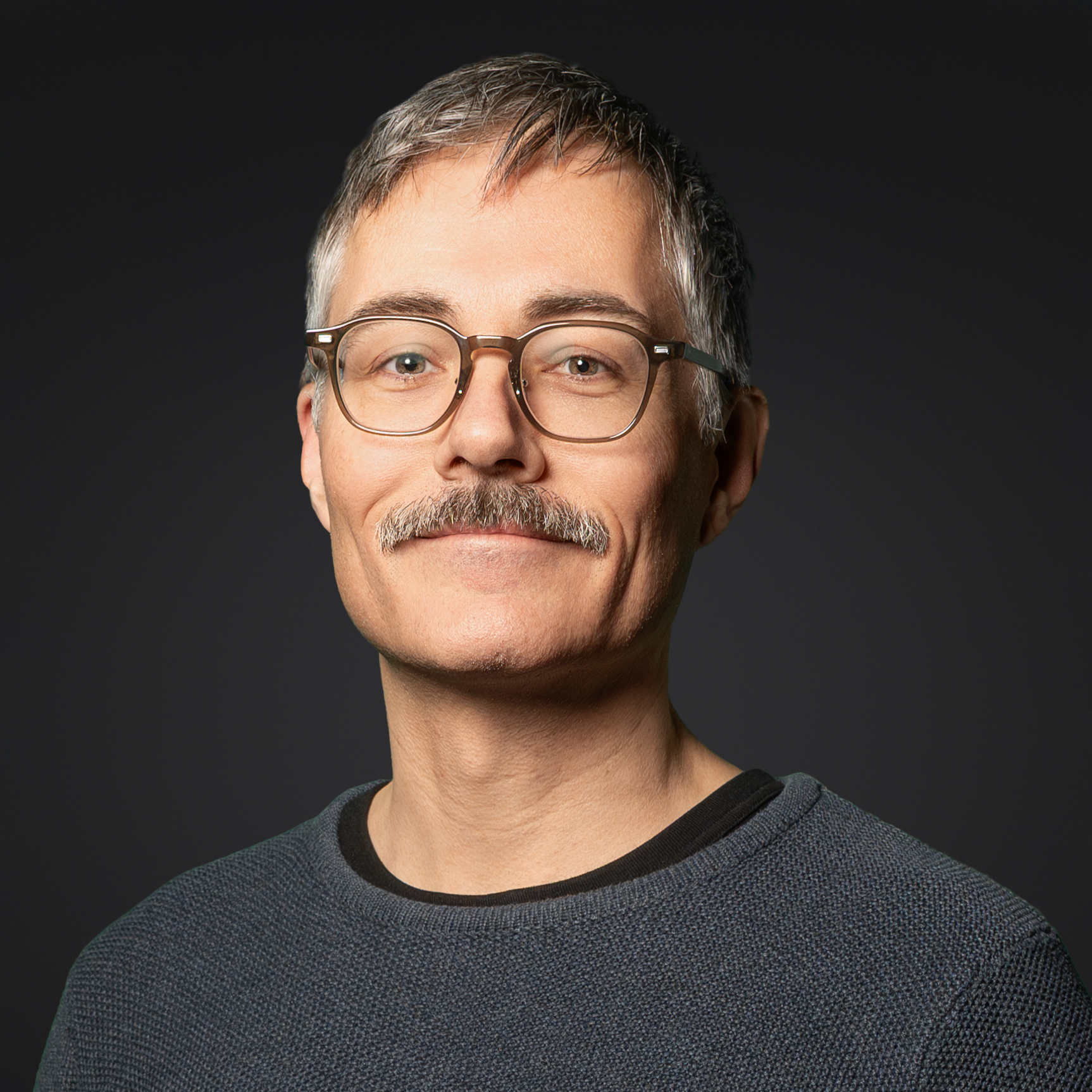 Oberkörperporträt eines Mannes mit grauen Haaren und Brille, der einen dunkelblauen Pullover über einem schwarzen T-Shirt trägt, vor dunklem Hintergrund.