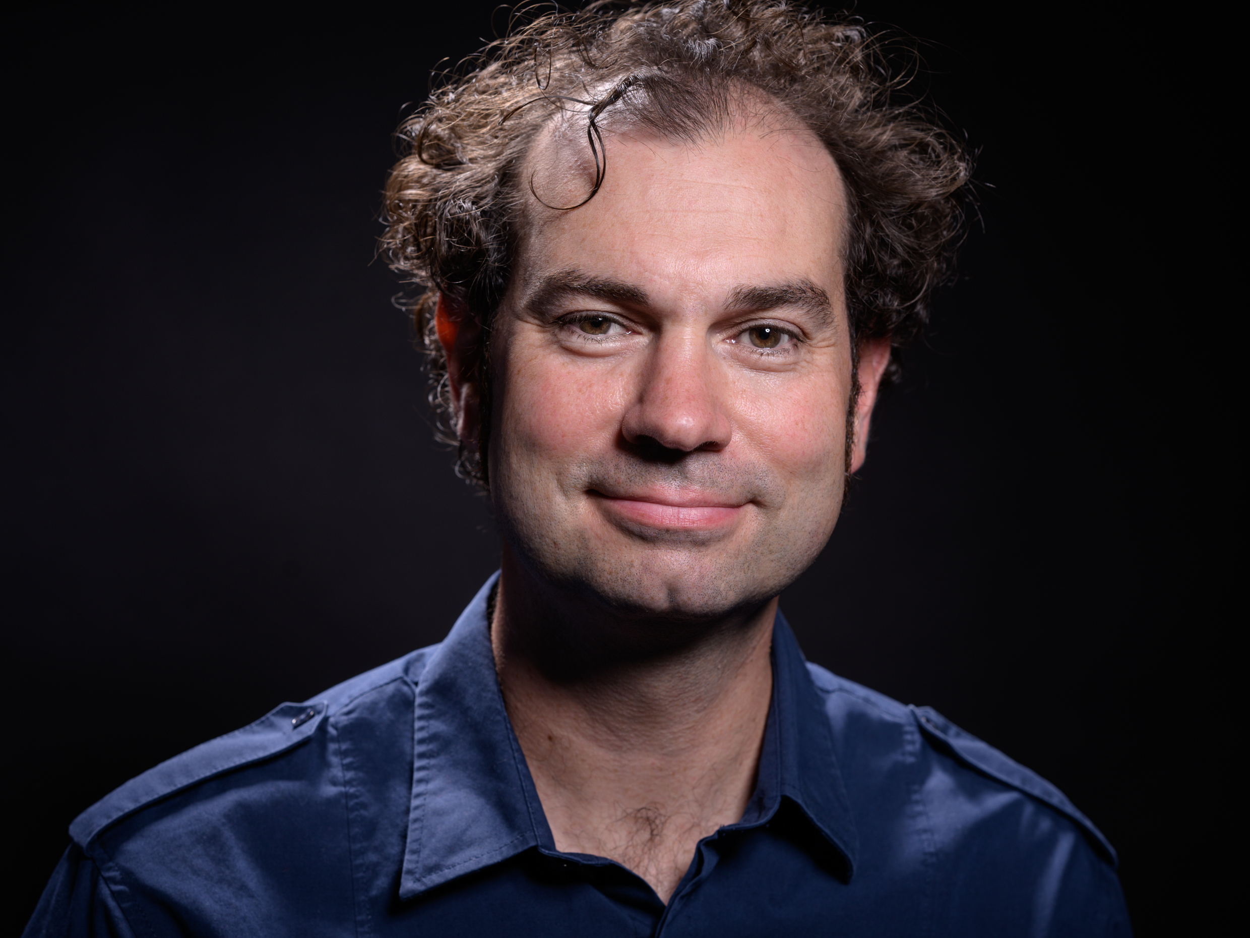 Mann mit lockigem Haar trägt ein dunkelblaues Hemd mit offenem Kragen vor schwarzem Hintergrund.