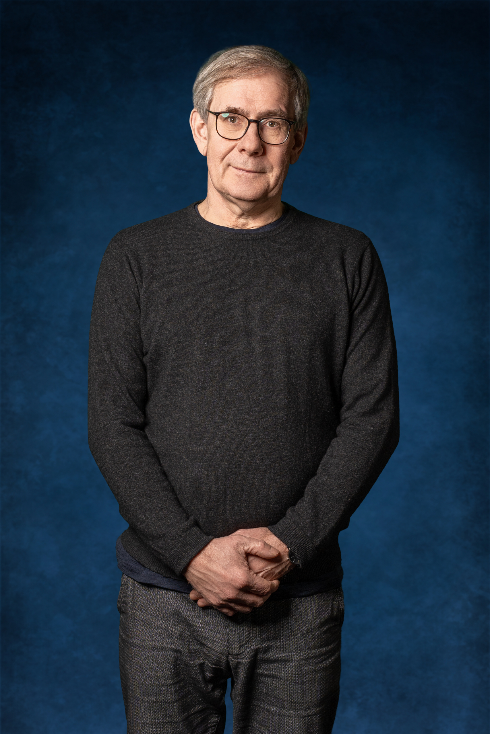 Mann mit Brille steht mit verschränkten Händen vorm Bauch in einem schwarzen Pullover vor einem blauen Hintergrund.