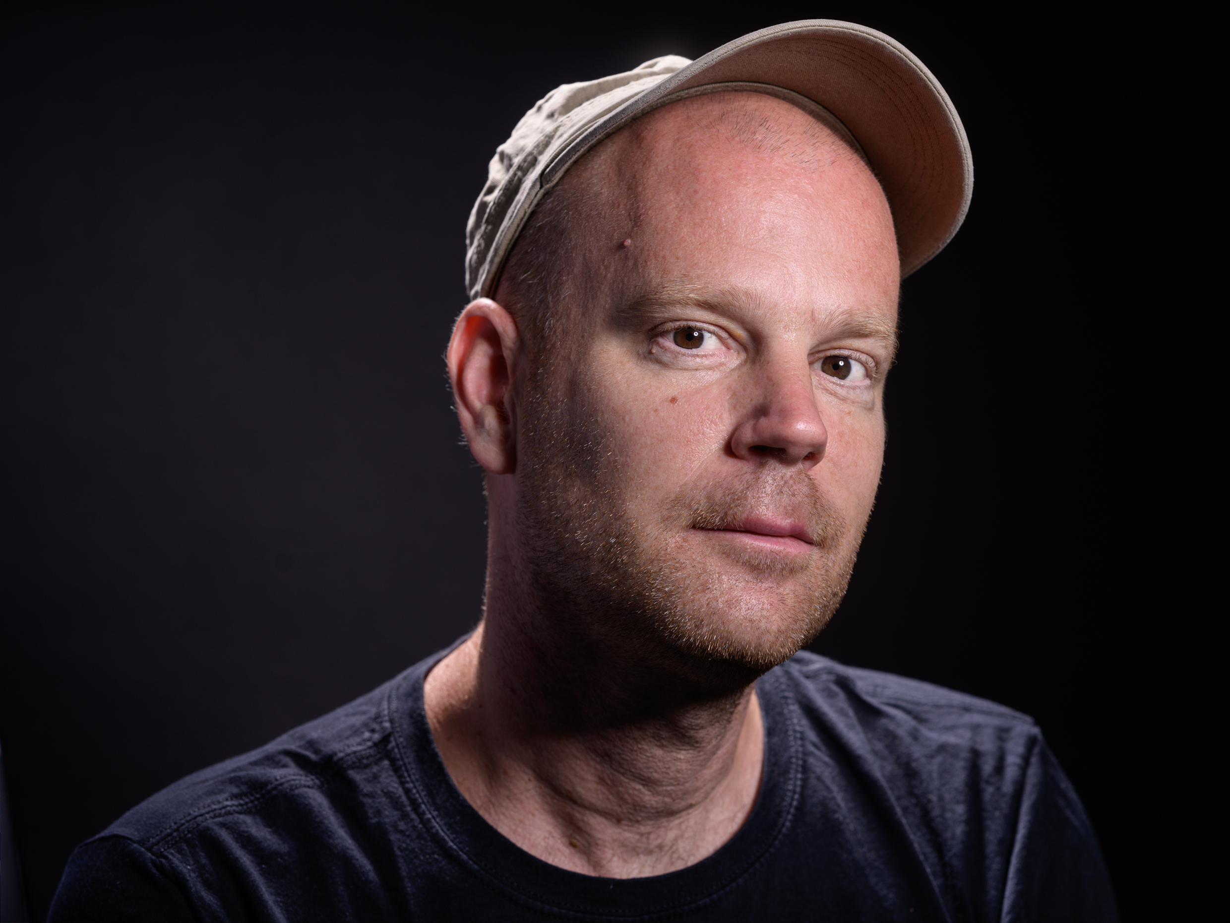 Mann mit hellbeiger Baseballkappe und dunkelblauem T-Shirt vor schwarzem Hintergrund.