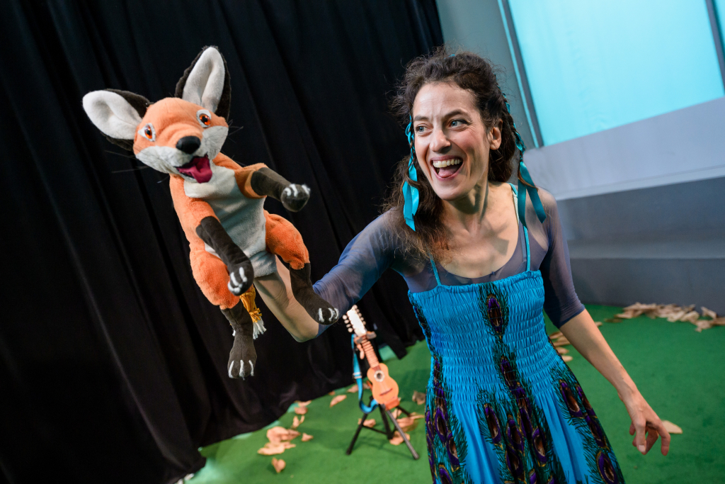 Frau in blauem Kleid hält eine orange-weiße Fuchs-Handpuppe mit offenem Mund und ausgestreckten Armen.