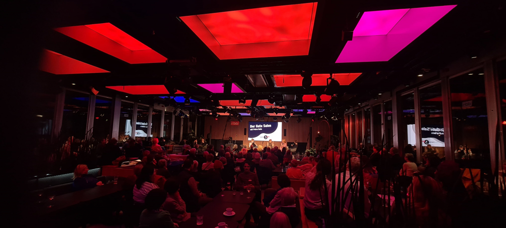 Großer Veranstaltungsraum mit Publikum vor einer Bühne und rot-violett beleuchtetem Deckenraster